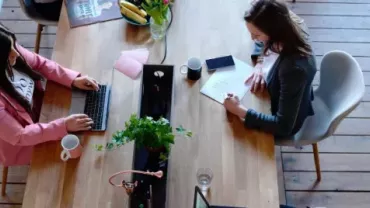 Zwei Personen arbeiten an einem Holztisch, Laptop und Notizen.